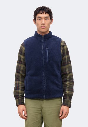 Un homme pose avec un gilet polaire bleu marine zippé sur une chemise à carreaux verts et un pantalon kaki, devant un fond clair et uni.