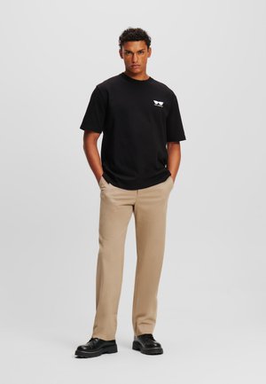 Black cotton t-shirt with short sleeves and a white graphic logo, paired with beige trousers and black shoes, featuring a smooth texture.