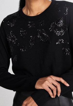 Czarny sweter z długim rękawem ozdobiony rozsianymi czarnymi cekinowymi kwiatowymi wzorami na piersiach i górnej części rękawów, noszony z rękami skrzyżowanymi na talii.