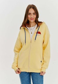 Žltá mikina na zips s kapucňou s hnedou podšívkou, modrými šnúrkami, prednými vreckami a vyšívaným detailom croissantu na ľavej hrudi.