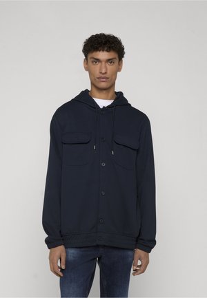 Homme portant un sweat à capuche bleu marine à boutons avec poches à rabat, debout devant un fond neutre, face à l'avant.