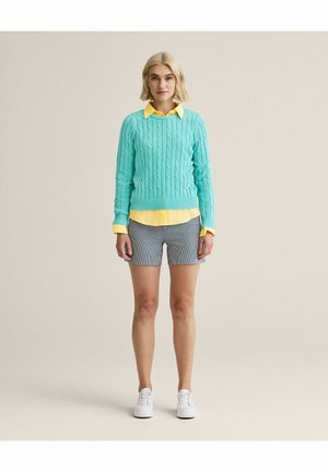 Kvinde stående iført en turkis kabelstrikket sweater over en gul skjorte, grå shorts og hvide sneakers mod en ensfarvet beige baggrund.