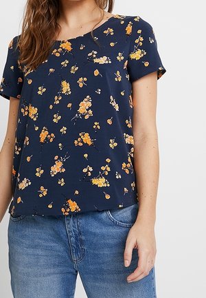 Marineblauwe korte mouwen blouse met geel en oranje bloemenprint, gedragen met blauwe spijkerbroek, getoond van nek tot heupen.