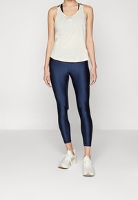 Canottiera atletica beige chiaro con accenti neri, abbinata a leggings blu scuro lucidi e sneakers bianche, in piedi su uno sfondo grigio.