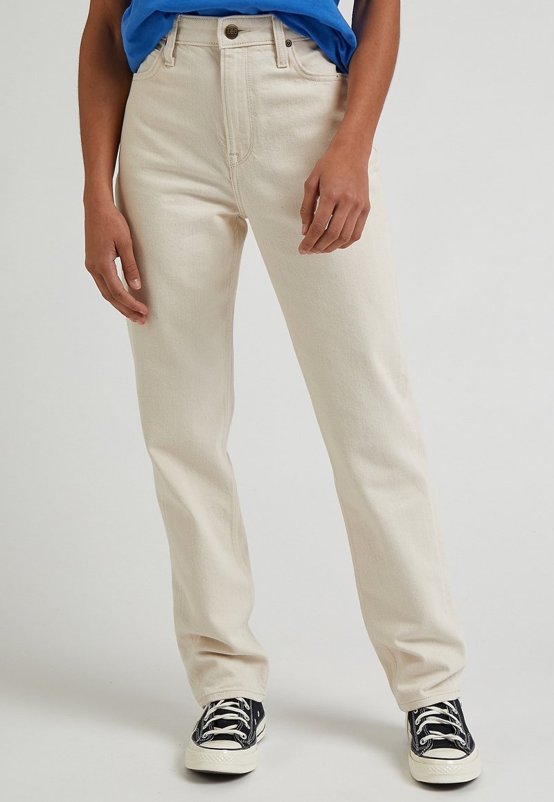 Béžové džíny rovného střihu vyrobené z denimu, s knoflíkovým zapínáním, poutky na opasek a předními kapsami, zkombinované s černými teniskami a modrým tričkem.