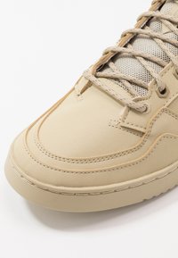 Béžové tenisky s hladkým syntetickým materiálem, texturovaným nápletem, tónovaným prošíváním a zpevněnými očky na šňůrky. Zaoblená gumová podrážka.