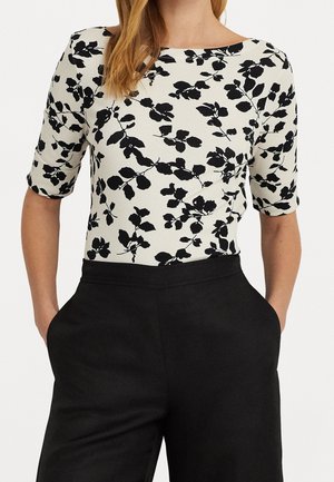 Kobieta w białej bluzce z czarnymi liściastymi wzorami i rękawami do łokci, w połączeniu z wysokim stanem czarnych spodni, trzymająca ręce w kieszeniach.