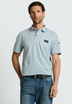 Mann mit kurzen lockigen Haaren, der ein hellblaues Polohemd mit gestreiftem Kragen und blaue Jeans trägt, vor einem weißen Hintergrund steht.