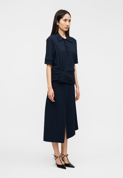 Το φόρεμα σε ναυτικό μπλε διαθέτει κοντομάνικο τοπ με γιακά και κουμπιά, με ζώνη στη μέση, που καταλήγει σε μια φούστα μέσης μήκους με ανοίγματα στο πλάι.