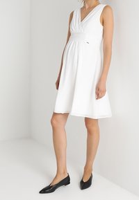 Vit ärmlös klänning med V-halsringning, rynkad midja och utsvängd kjol. Kombinerad med svarta spetsiga ballerinaskor mot en slät, neutral bakgrund.