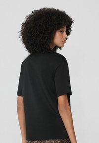 Czarna koszulka z krótkim rękawem, z okrągłym dekoltem. Gładki materiał, luźny krój, ukazująca widok z tyłu i subtelne detale strukturalne na powierzchni.