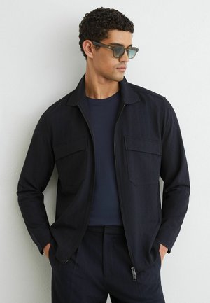 Homme portant une veste zippée bleu marine foncé sur une chemise bleu marine et un pantalon assorti, avec des lunettes de soleil, debout contre un mur blanc, les mains dans les poches.