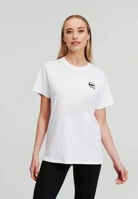 White cotton t-shirt with short sleeves and a round neck. Features a small black graphic on the left chest area. Straight-cut design.