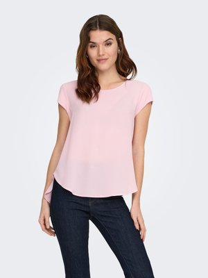 Mujer que lleva una blusa de manga corta color rosa claro y jeans azul oscuro, de pie contra un fondo liso gris claro.