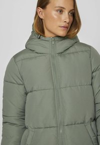 Veste matelassée vert clair avec une capuche, dotée de coutures horizontales, d'une fermeture éclair à l'avant et de deux poches latérales. Texture lisse et mate.