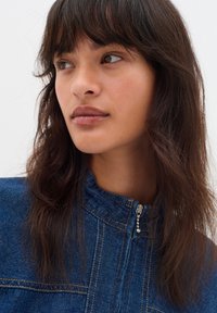 Jeansjacke mit hohem Kragen, blauer Farbe, goldenen Nähten und einem silbernen Reißverschluss mit dekorativen Perlen. Schlichte Gestaltung.