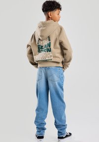Sudadera beige con estampado gráfico en la parte posterior, combinada con pantalones vaqueros sueltos de color azul claro y zapatillas negras. La tela parece suave con un corte relajado.