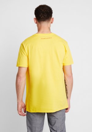 Mężczyzna z krótkimi kręconymi włosami, ubrany w jasny żółty t-shirt z napisem "NIGHT ADDICT" na górnej części pleców oraz szare spodnie w kratę.