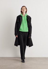 Impermeabil negru peste un hanorac verde aprins, asociat cu pantaloni negri și pantofi negri. Material neted cu un design lejer.