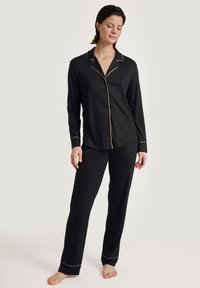 Zwarte pyjama set met een langelijnafgeknopte top en rechte broekspijpen. Gouden piping accenten op de kraag, manchetten en zoom. Zacht textuur van de stof.