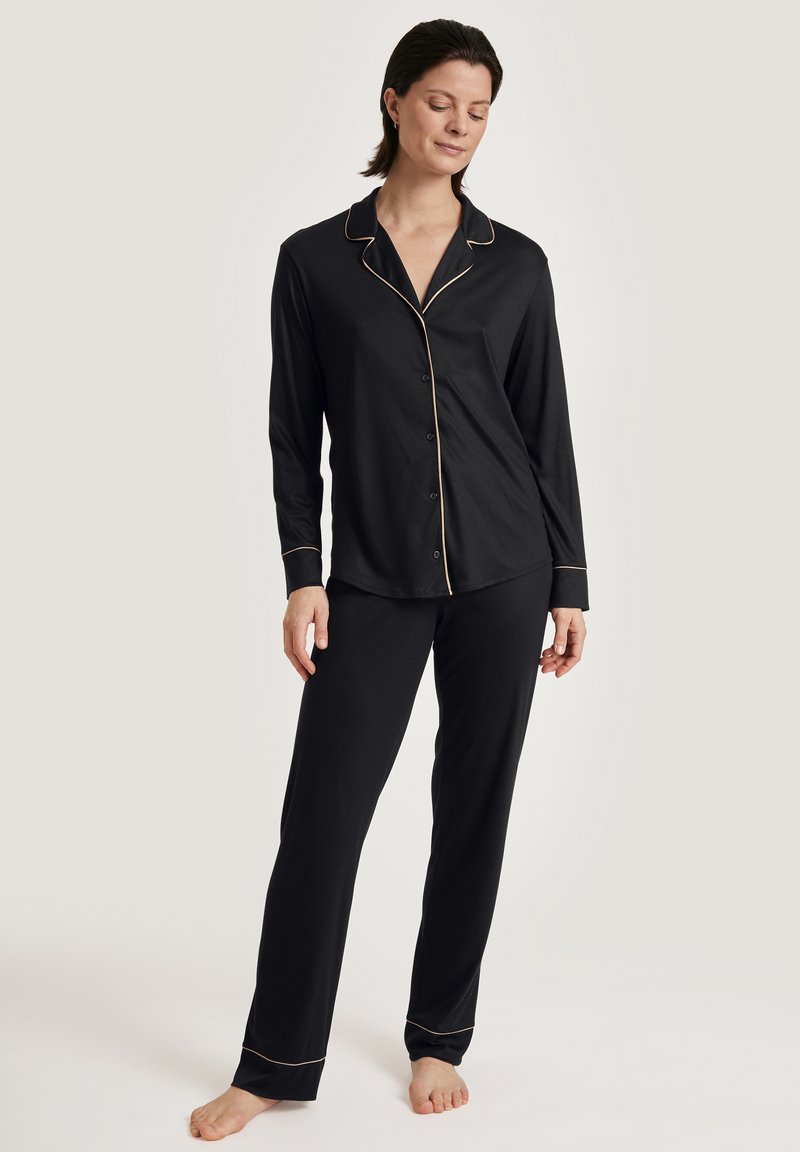 Zwarte pyjama set met een langelijnafgeknopte top en rechte broekspijpen. Gouden piping accenten op de kraag, manchetten en zoom. Zacht textuur van de stof.