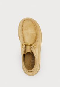 Beigās zamšādas kurpes ar noapaļotu formu. Iekļauti divi acu caurumi šņorēšanai un izšūti rotājumi apkārt augšdaļai. Tekstūrēts iekšējais vožs.