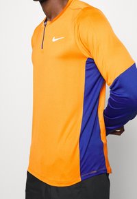 Orange og lilla langærmet atletisk skjorte med kvart-zip krave, der har et struktureret stof og et lille hvidt logo på brystet.