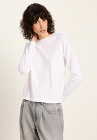 Frau mit lockigem dunklem Haar, die ein weißes Langarmshirt und hellgraue Jeans trägt, posiert mit einer Hand hinter ihrem Nacken.