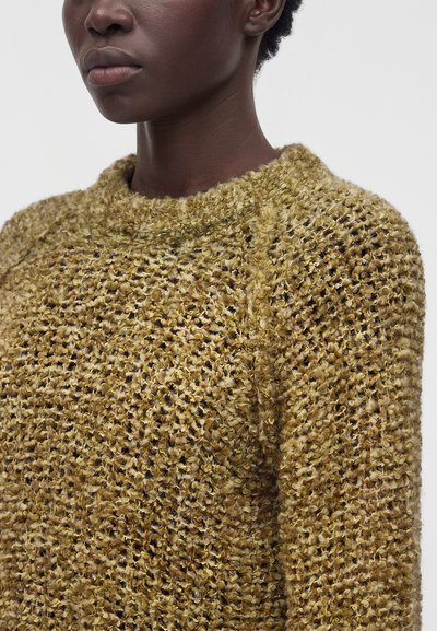 Gros plan d'une personne portant un pull en maille jaune moutarde texturé avec un col rond sur un fond uni.