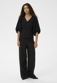 Melnas teksturētas blūzes ar V-veida kakla izgriezumu un pūkainām piedurknēm, apvienotas ar augsto vidukli melnām platām biksēm un ar smailiem melniem apaviem.