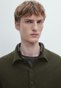 Zielony sweter z teksturowanym splotem, z kołnierzykiem i trzema guzikami, nałożony na ciemnoszary t-shirt. Skupienie na górnej części ciała i dekolcie.