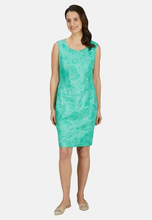 Femme debout dans une robe turquoise sans manches avec des motifs cachemire, portant des chaussures plates beiges, souriante, les cheveux tombant sur une épaule.