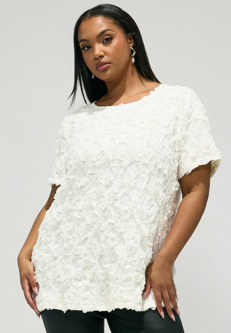 Femme portant un haut blanc texturé à manches courtes et motifs floraux, avec un col rond, associée à un bas noir, posant devant un fond uni.