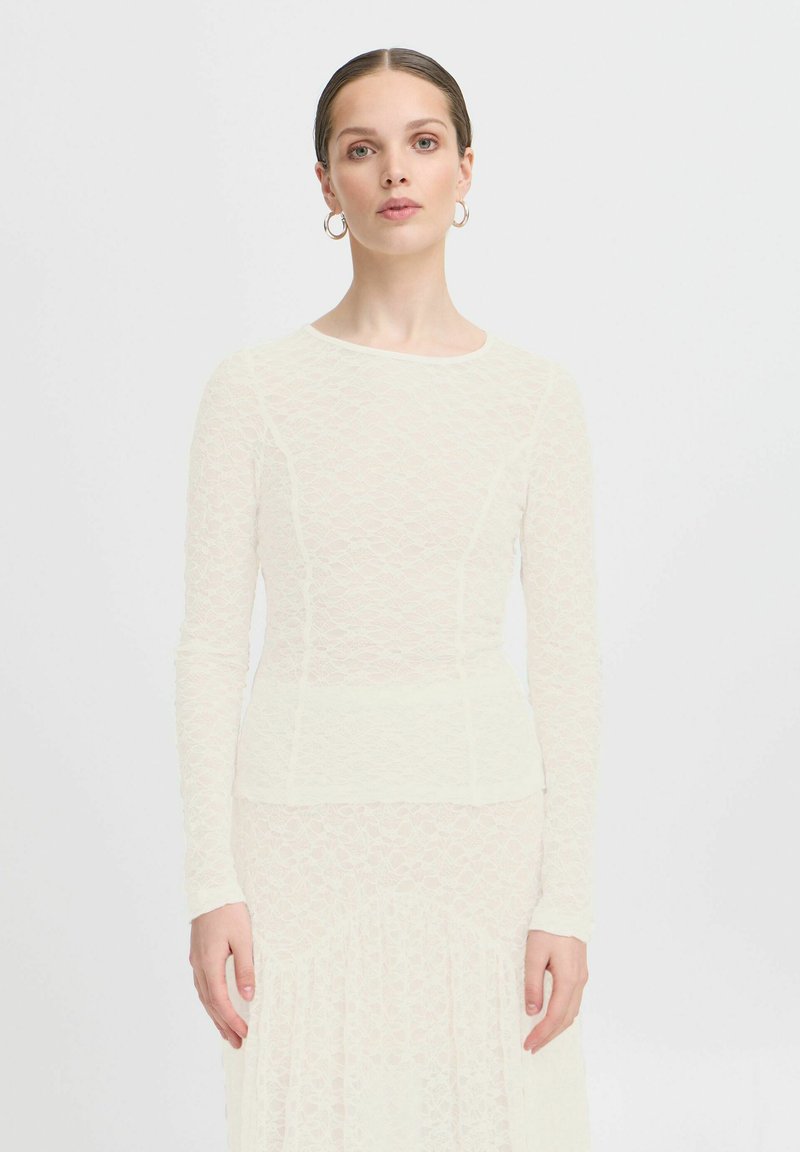 Top in pizzo avorio a maniche lunghe con un design aderente, scollatura rotonda e motivo testurizzato, sovrapposto a una parte inferiore fluida.