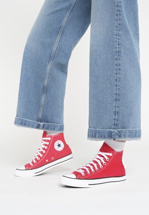 Ben i lyseblå vidde jeans iført knaldrøde høje Converse-sneakers med hvide snørebånd og såler på en hvid baggrund.