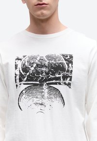 Artimos planas baltos, ilgomis rankovėmis marškinėlių su juodu abstrakčiu apskritu atspaudu, primenančiu medžio žiedus, ant krūtinės.