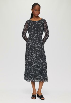 Schwarzes Kleid mit langen durchsichtigen Ärmeln, floralen weißen Muster, taillierter Oberteil, plissierter Taillenbund und gewelltem Saum. Getragen mit schwarzen Riemchenschuhen.