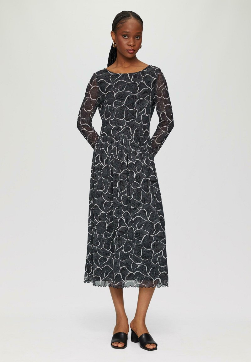 Schwarzes Kleid mit langen durchsichtigen Ärmeln, floralen weißen Muster, taillierter Oberteil, plissierter Taillenbund und gewelltem Saum. Getragen mit schwarzen Riemchenschuhen.