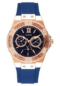 Roséguld klocka med marinblå silikonrem. Har en glittrande bezel, mörk urtavla med gulddetaljer och dubbla subdials för datum och tid.