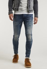 Jeans de mezclilla azul ajustados con áreas desgastadas y detalles rasgados, combinados con zapatos de suede beige y un sudadera gris bajo una chaqueta de mezclilla.