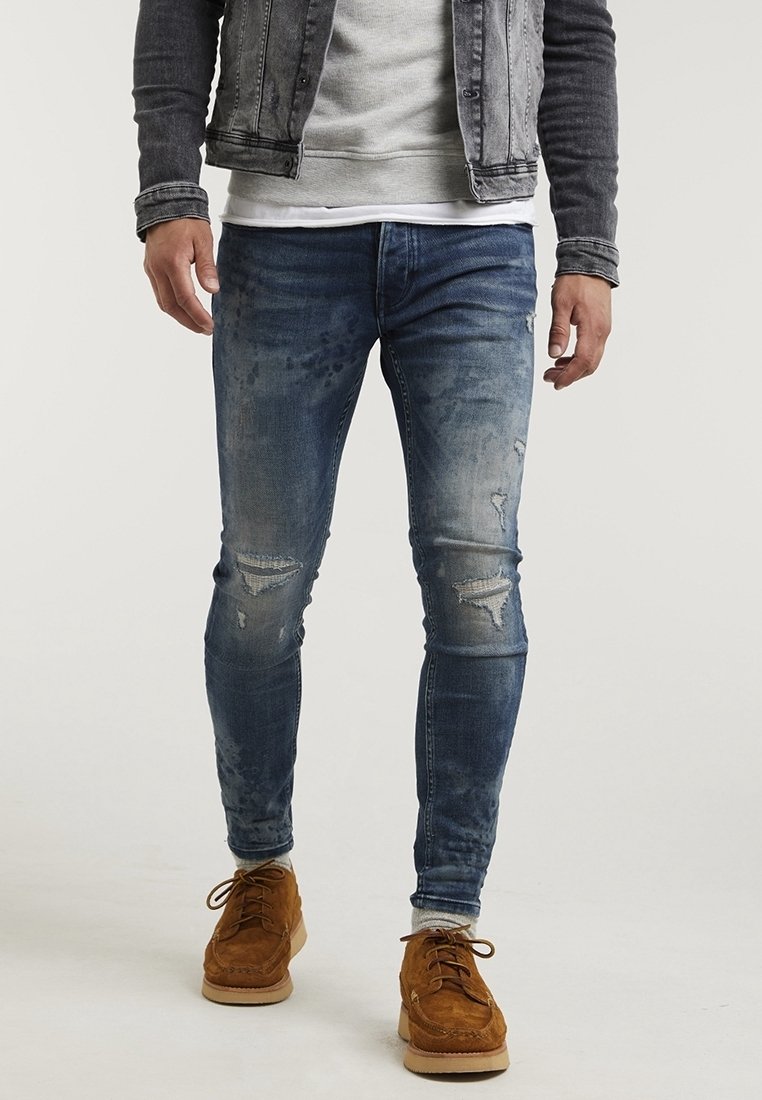 Jeans de mezclilla azul ajustados con áreas desgastadas y detalles rasgados, combinados con zapatos de suede beige y un sudadera gris bajo una chaqueta de mezclilla.