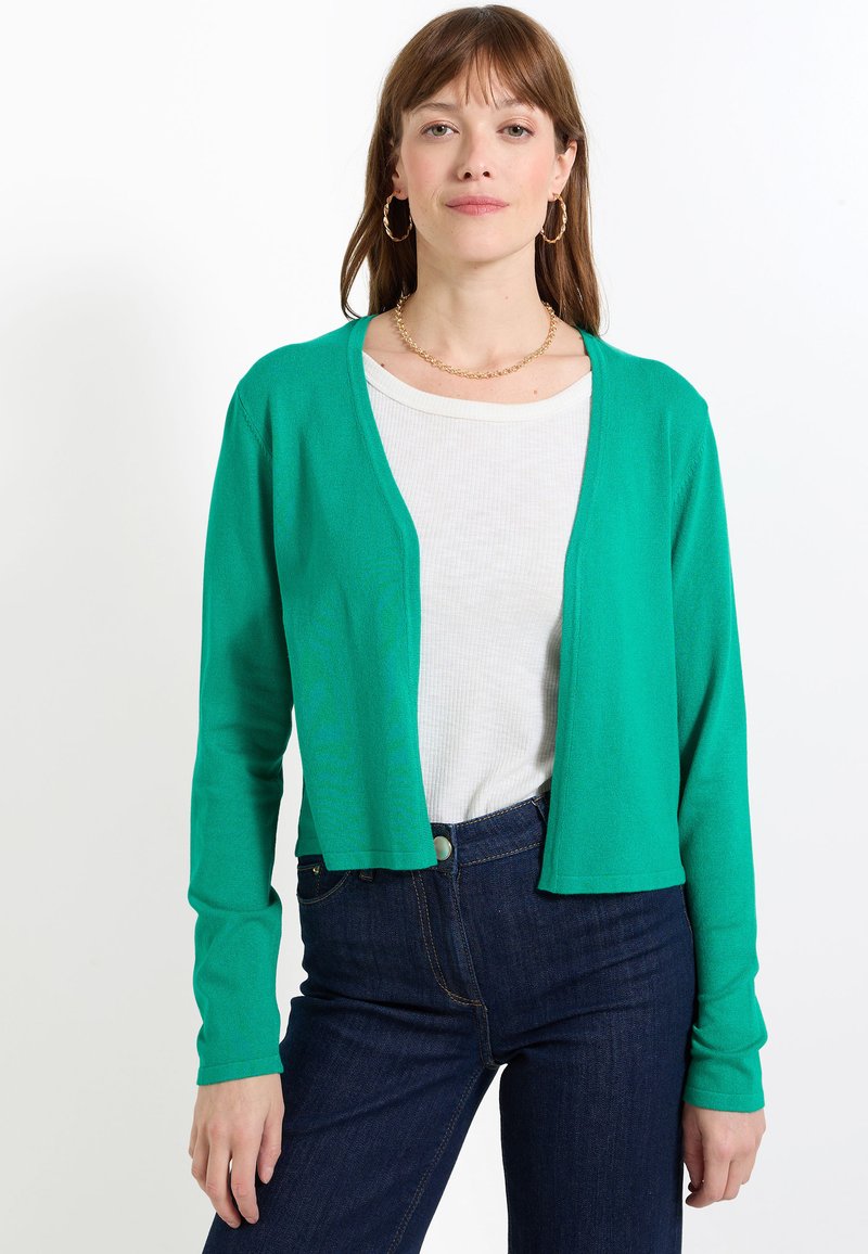 Cárdigan verde corto hecho de material ligero, con mangas largas y diseño de apertura frontal, usado sobre una blusa blanca y jeans oscuros.