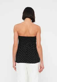 Svart stroppeløs topp med hvite polkadotter, med smocket overdel og et flytende design, vist bakfra mot en ensfarget bakgrunn.