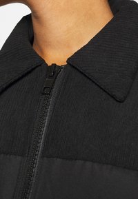 Gros plan sur le col d'une veste noire côtelée avec une fermeture éclair noire visible, portée par une personne à la peau de ton moyen.