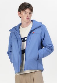 Jachetă cu fermoar de culoare albastru deschis, cu glugă, având un logo pe piept și interior căptușit cu fleece. Purtați-o peste un pulover crem cu dungi întunecate.
