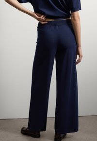 Persoon in marineblauwe wijde broek en bijpassende top, staand met één hand op de onderrug tegen een effen witte muur.