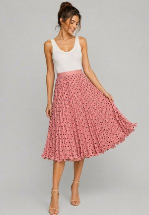 Femme avec les cheveux en chignon, portant un top blanc sans manches et une jupe midi plissée rose à pois noirs, tenant l'ourlet de la jupe, souriante.