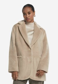 Beige Wollmischungsmantel mit Reverskragen, zwei Knöpfen und Fronttaschen. Weiche Textur und lockere Passform, kombiniert mit einem cremefarbenen Rollkragenpullover.