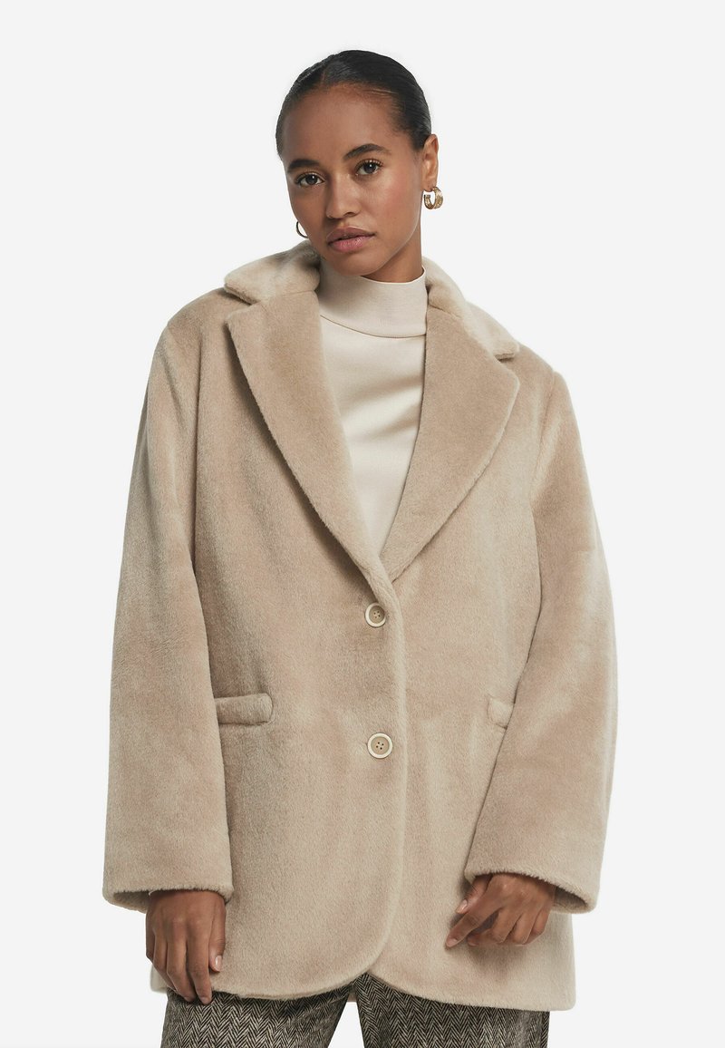 Beige Wollmischungsmantel mit Reverskragen, zwei Knöpfen und Fronttaschen. Weiche Textur und lockere Passform, kombiniert mit einem cremefarbenen Rollkragenpullover.
