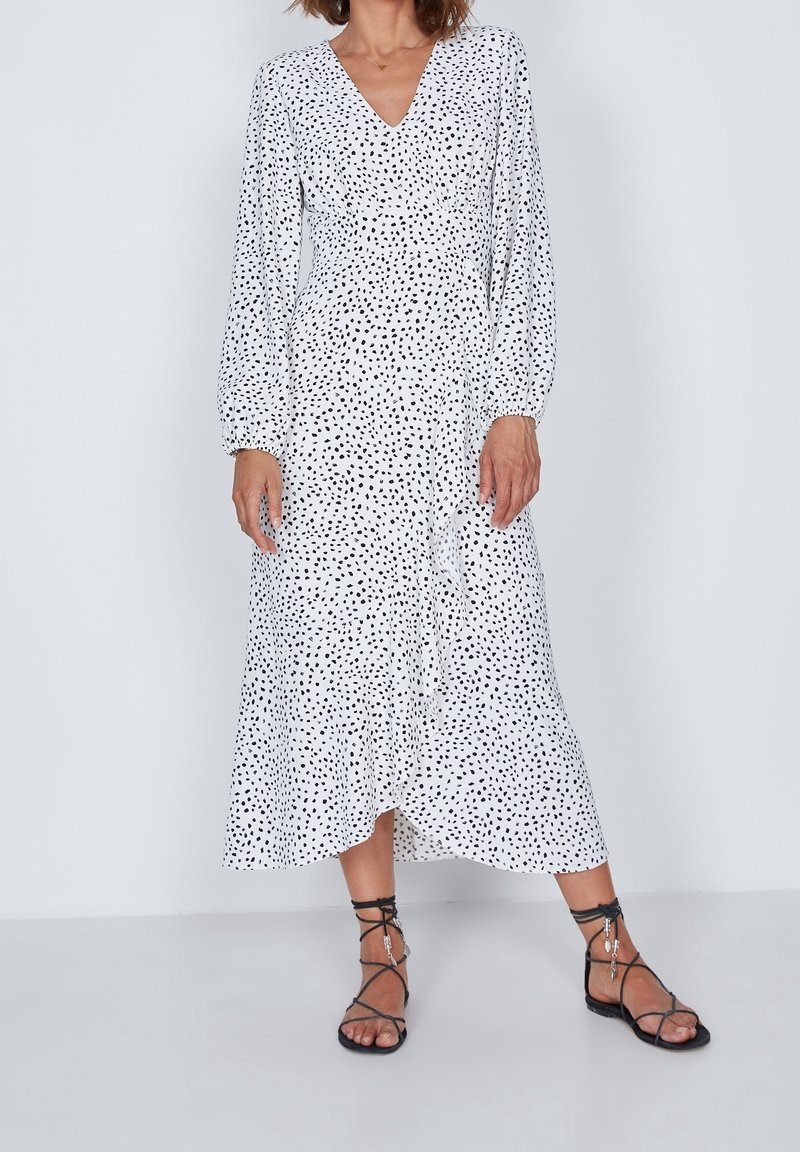Largo vestido blanco con lunares negros, escote en V, cintura fruncida y mangas largas abullonadas. Combinado con sandalias negras de tiras.